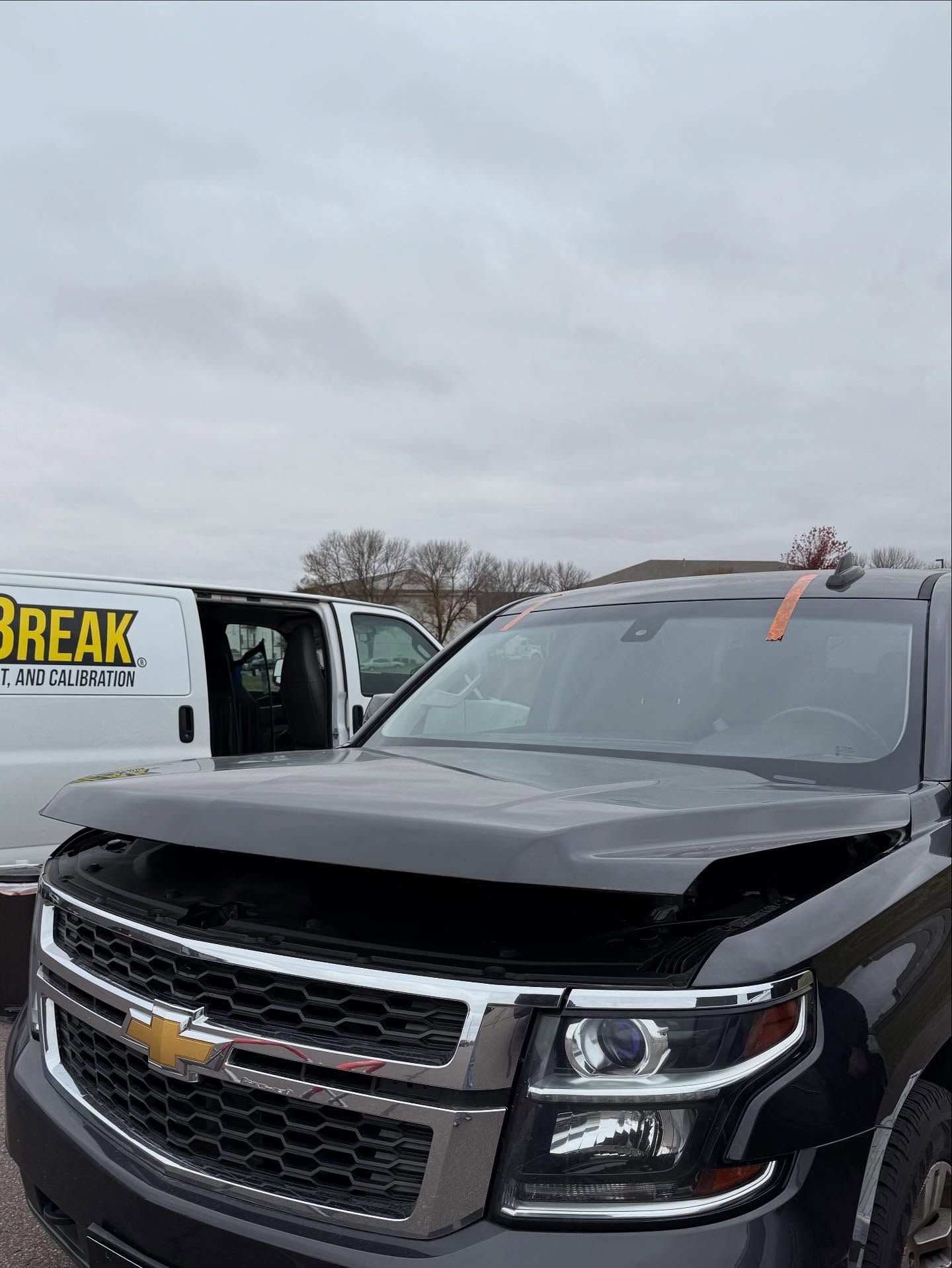 A dark gray Chevrolet SUV with its hood popped open parked in front of a white utility van with lettering.