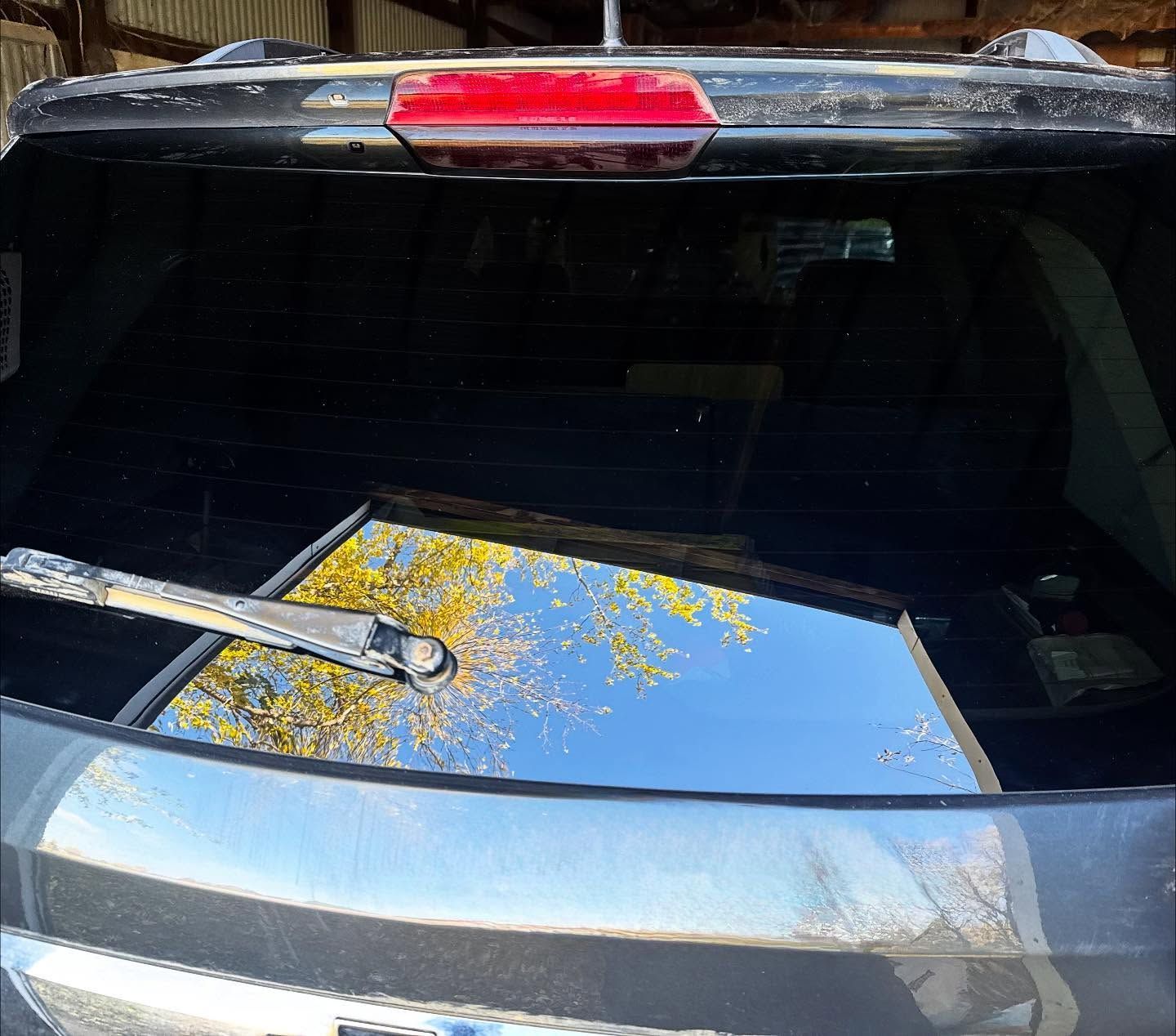 A dark gray car's rear windshield, featuring a horizontal wiper and a reflection of yellow autumn trees and blue sky.