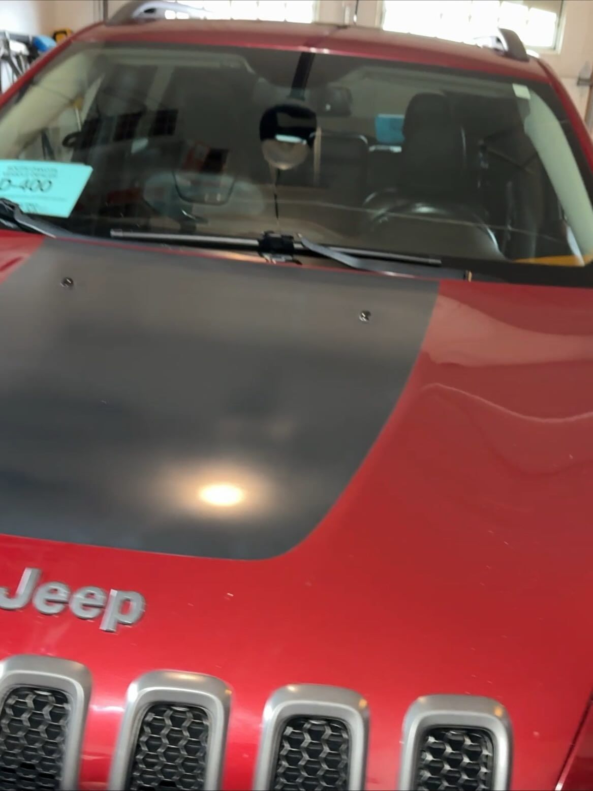 Close-up of a red Jeep with a matte black hood panel, showing the grille, hood, and windshield inside a garage.