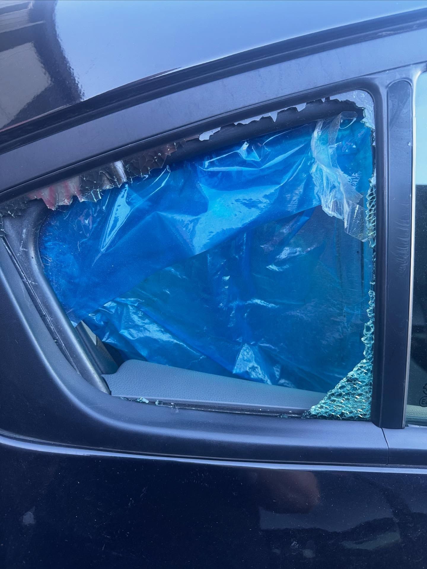 A car's rear passenger window is shattered, with a blue plastic sheet temporarily covering the opening.