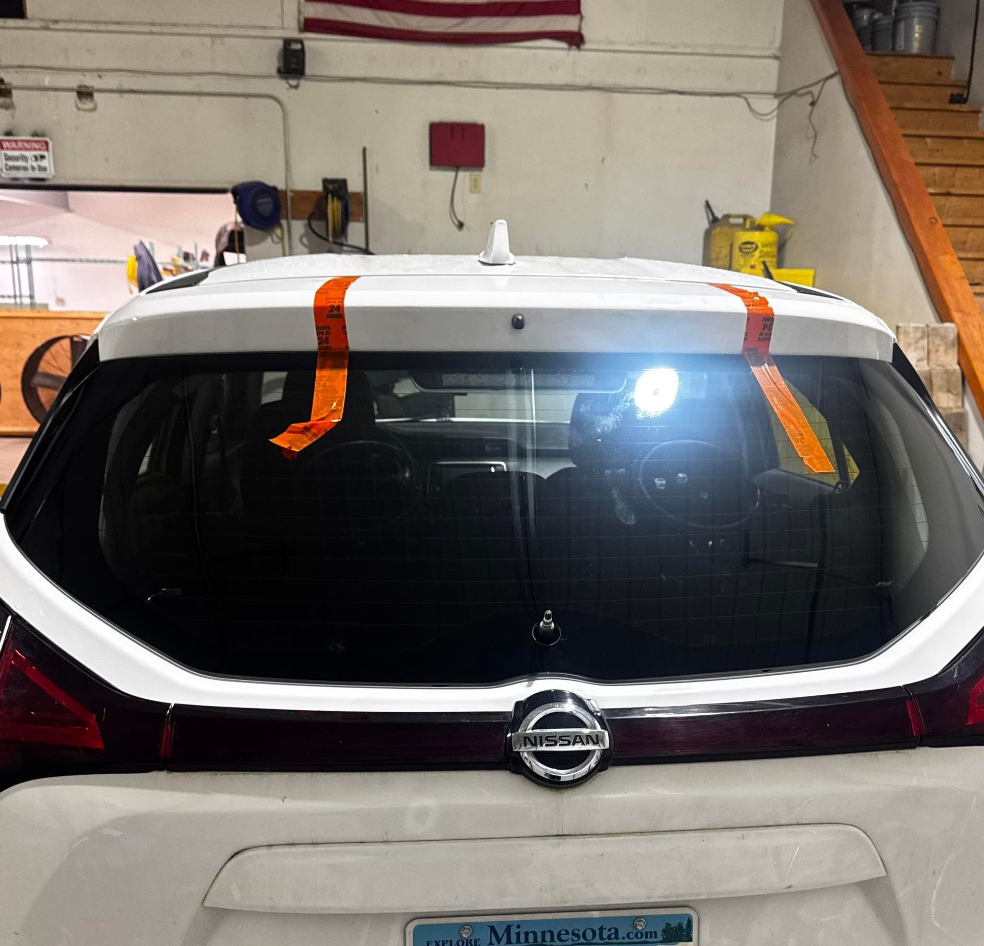 A rear view of a white Nissan hatchback inside a workshop, with orange tape strips holding the top spoiler in place.