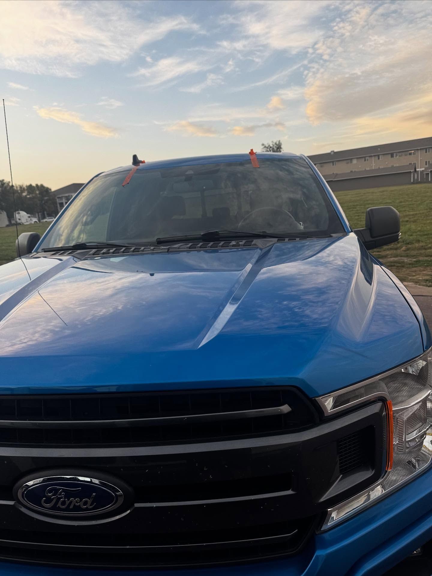 A blue Ford truck parked outdoors under a partially cloudy sky, focusing on the hood, grille, and windshield.