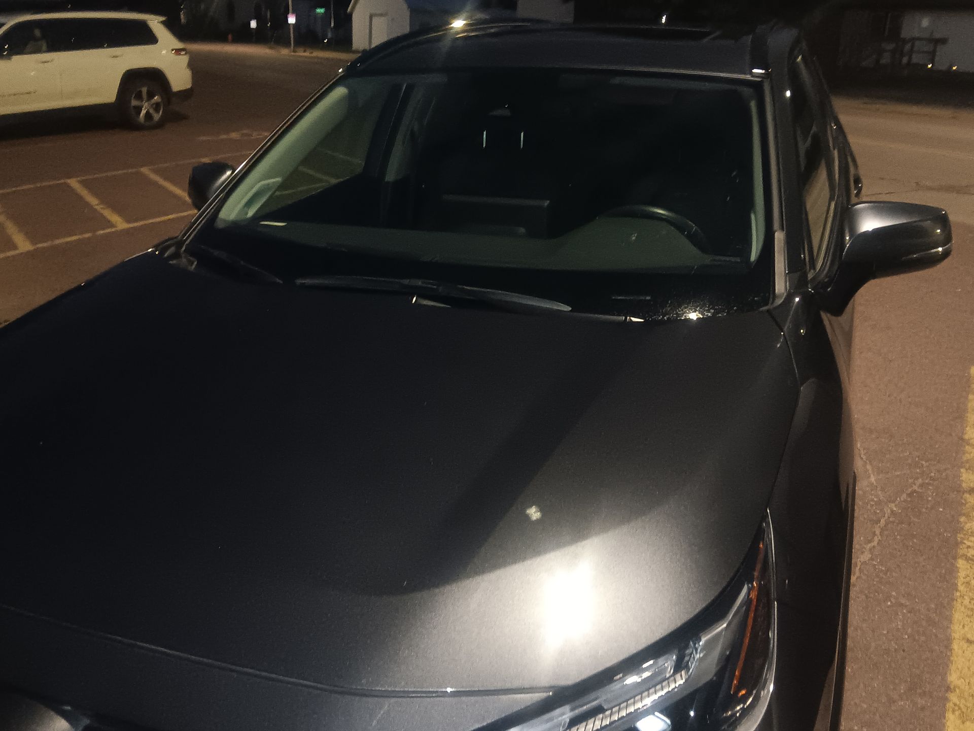 A dark gray SUV parked in a paved lot at night, with a white vehicle visible in the background.