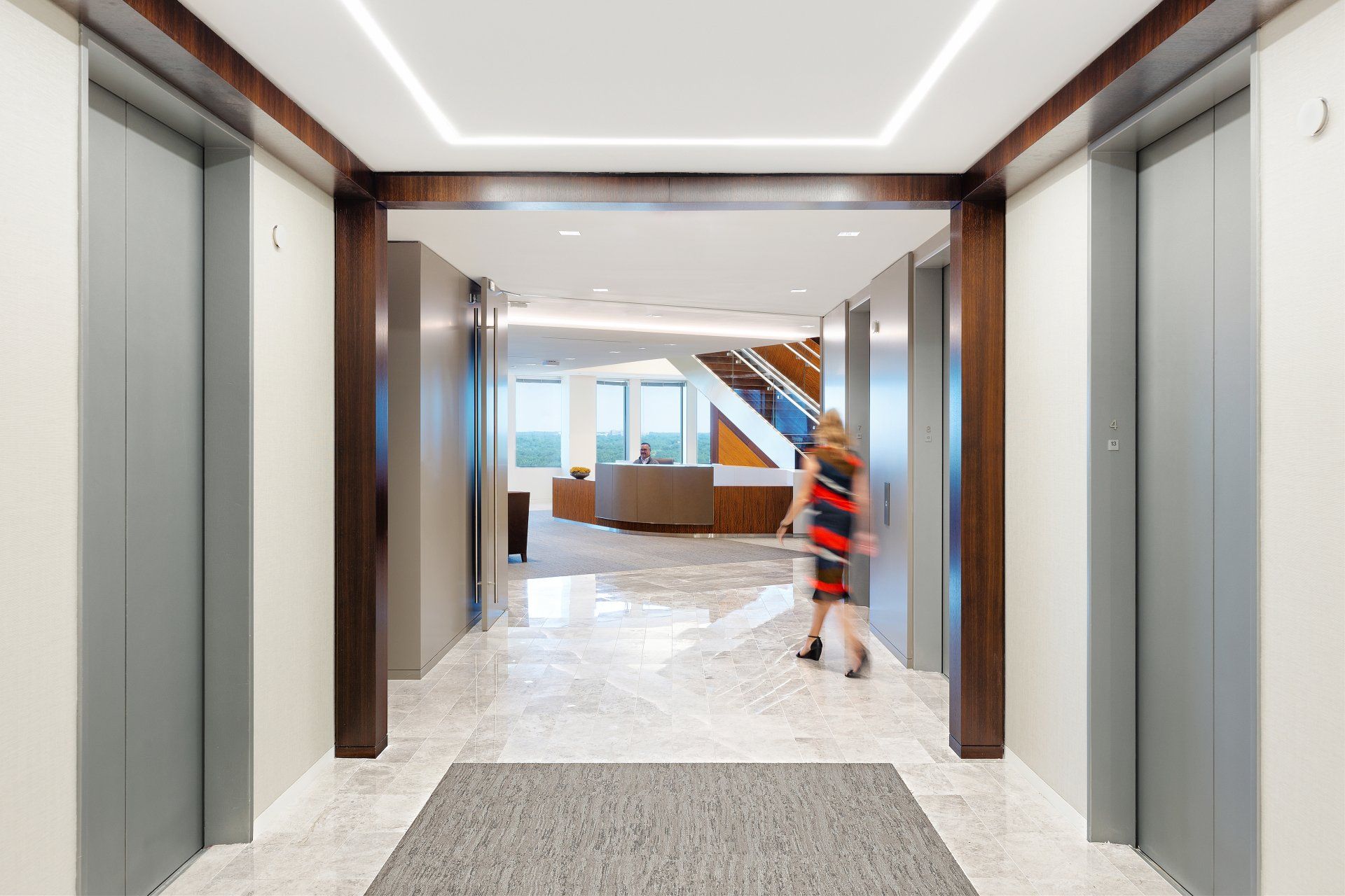 A woman is walking down a hallway between two elevators in an office building.