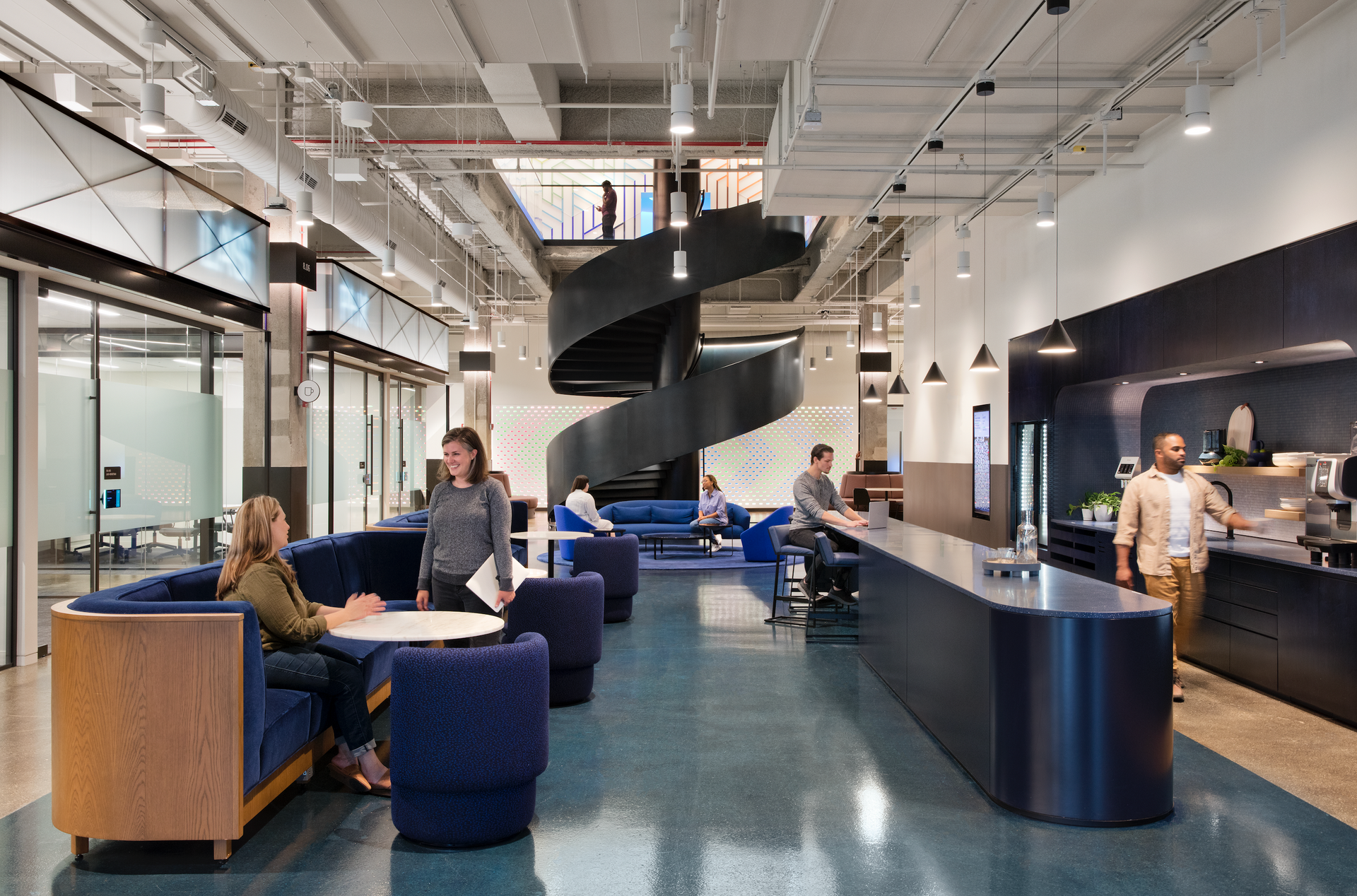 A group of people are sitting at tables in a large room with a spiral staircase.