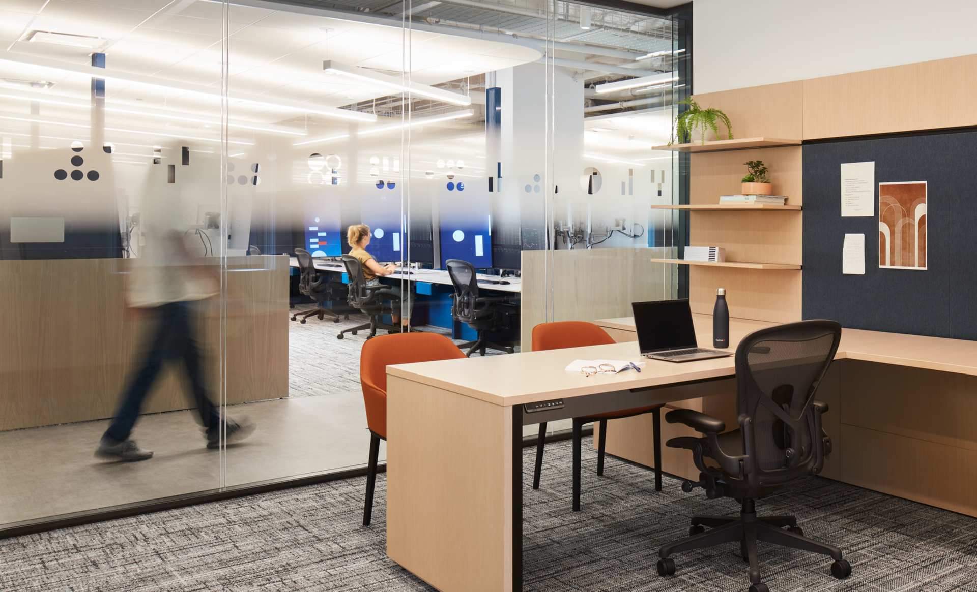 A man is walking past a desk in an office with a laptop on it.