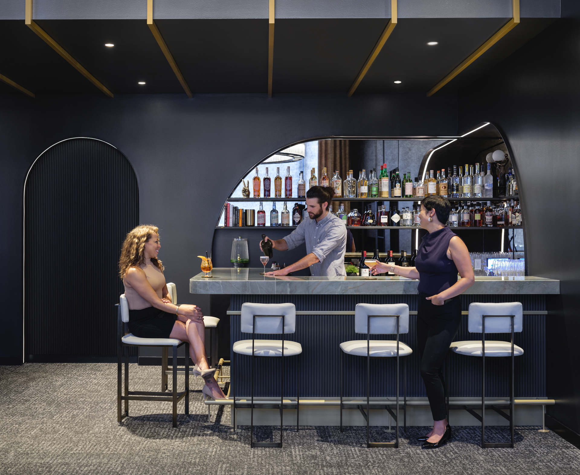 A man is pouring a drink into a glass while two women sit at a bar.