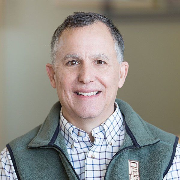 A man wearing a plaid shirt and a green vest is smiling for the camera.