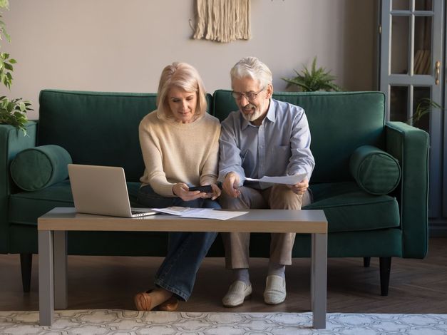 An elderly couple is sitting on a couch looking at a laptop.