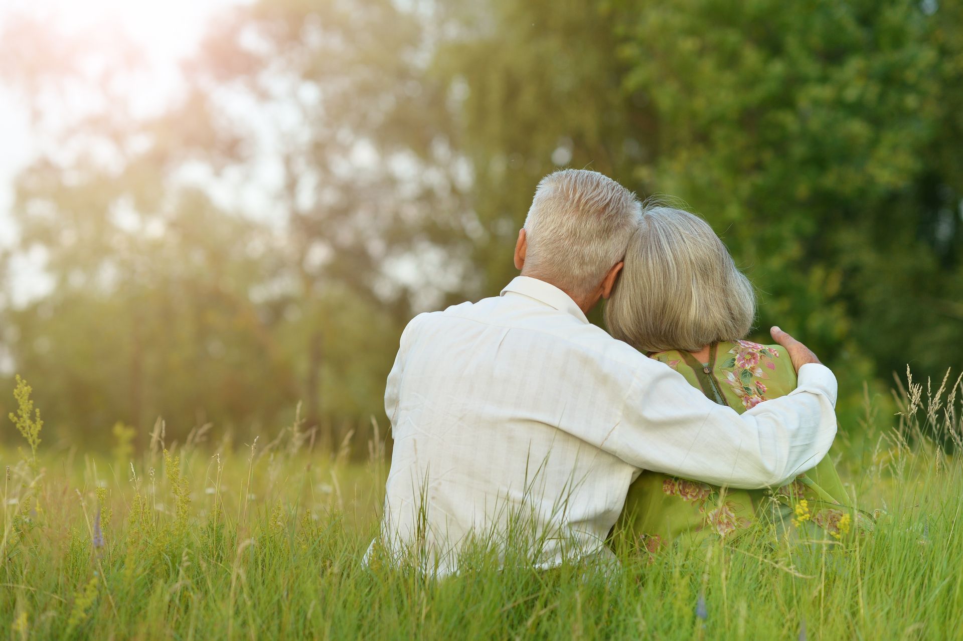 An elderly couple is sitting in a field hugging each other.