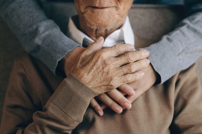 An elderly man is sitting in a chair with his hands on his chest.