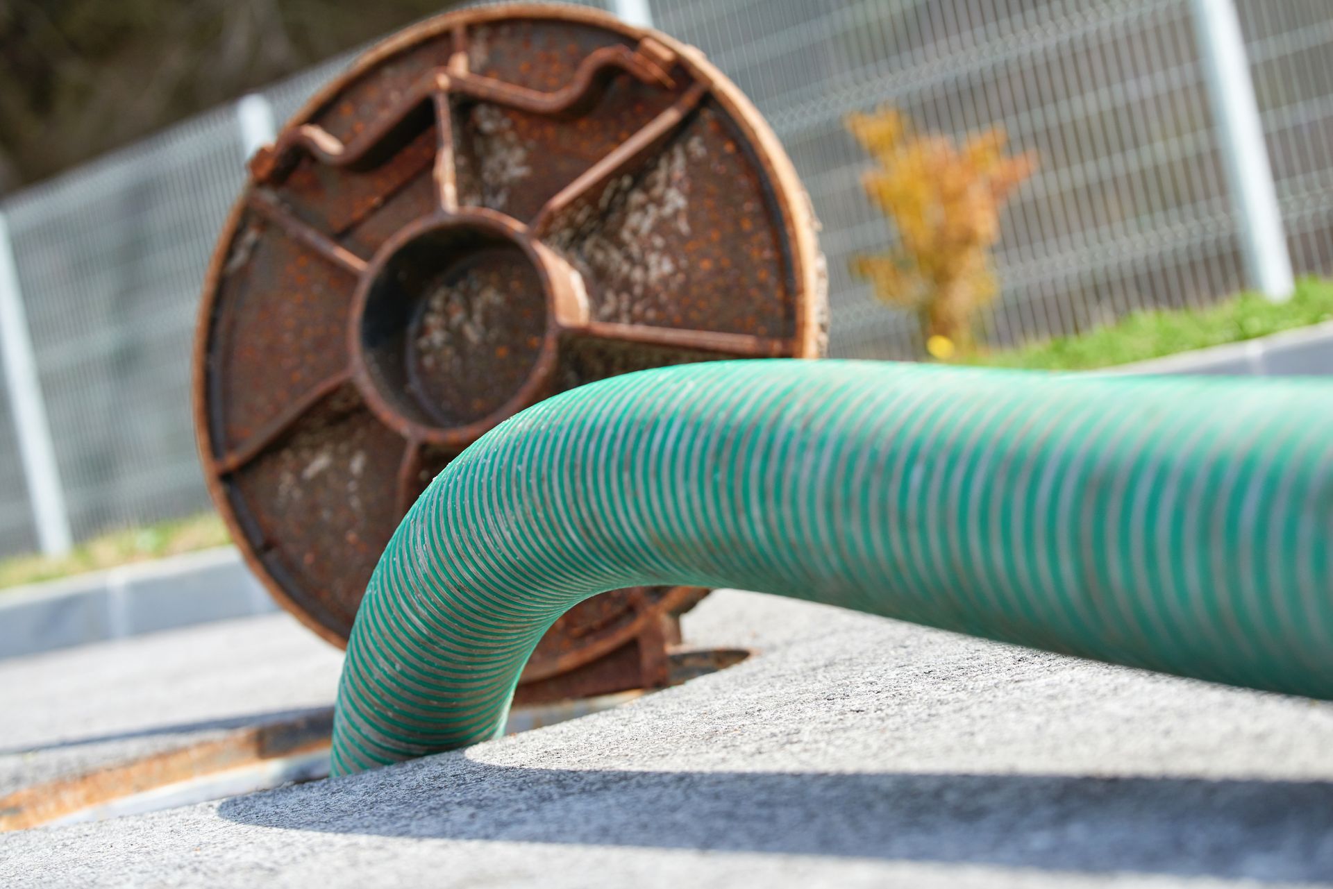 Green corrugated hose entering ground beside rusted metal reel near fence.