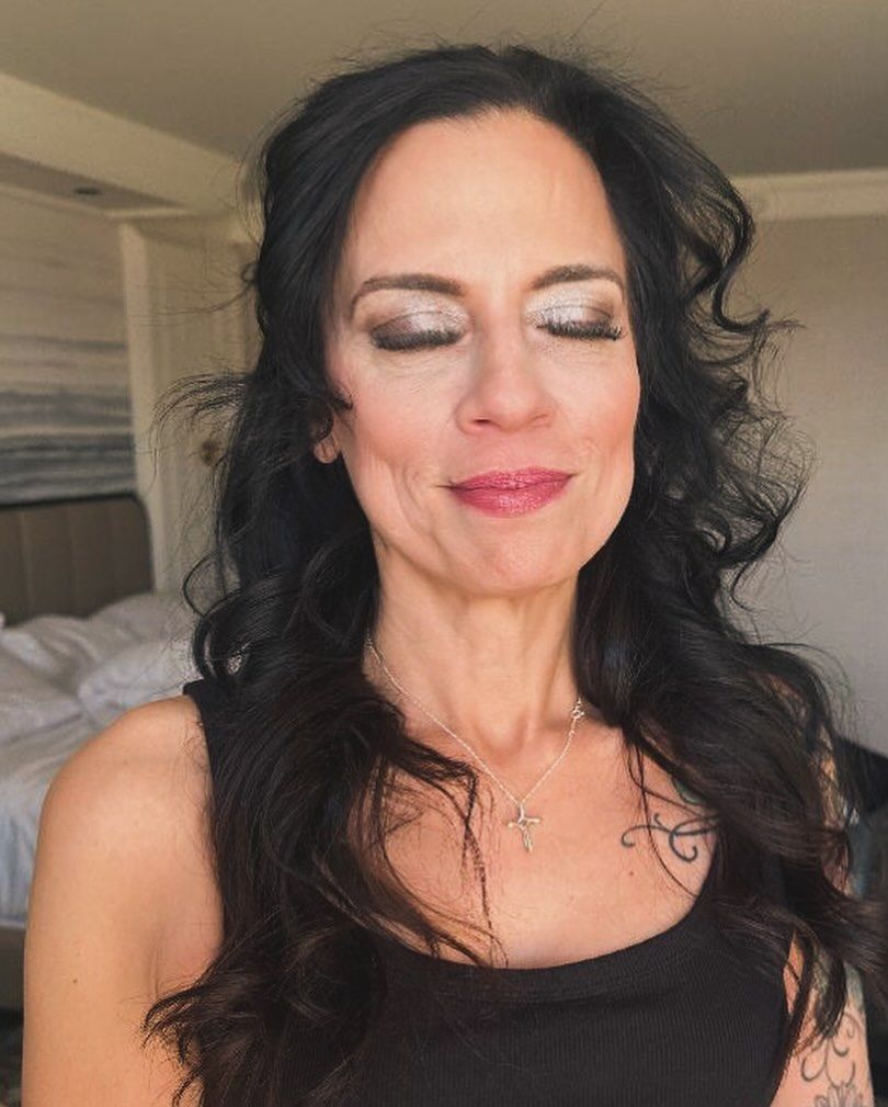 A woman with long black hair is wearing a black tank top and a cross necklace.