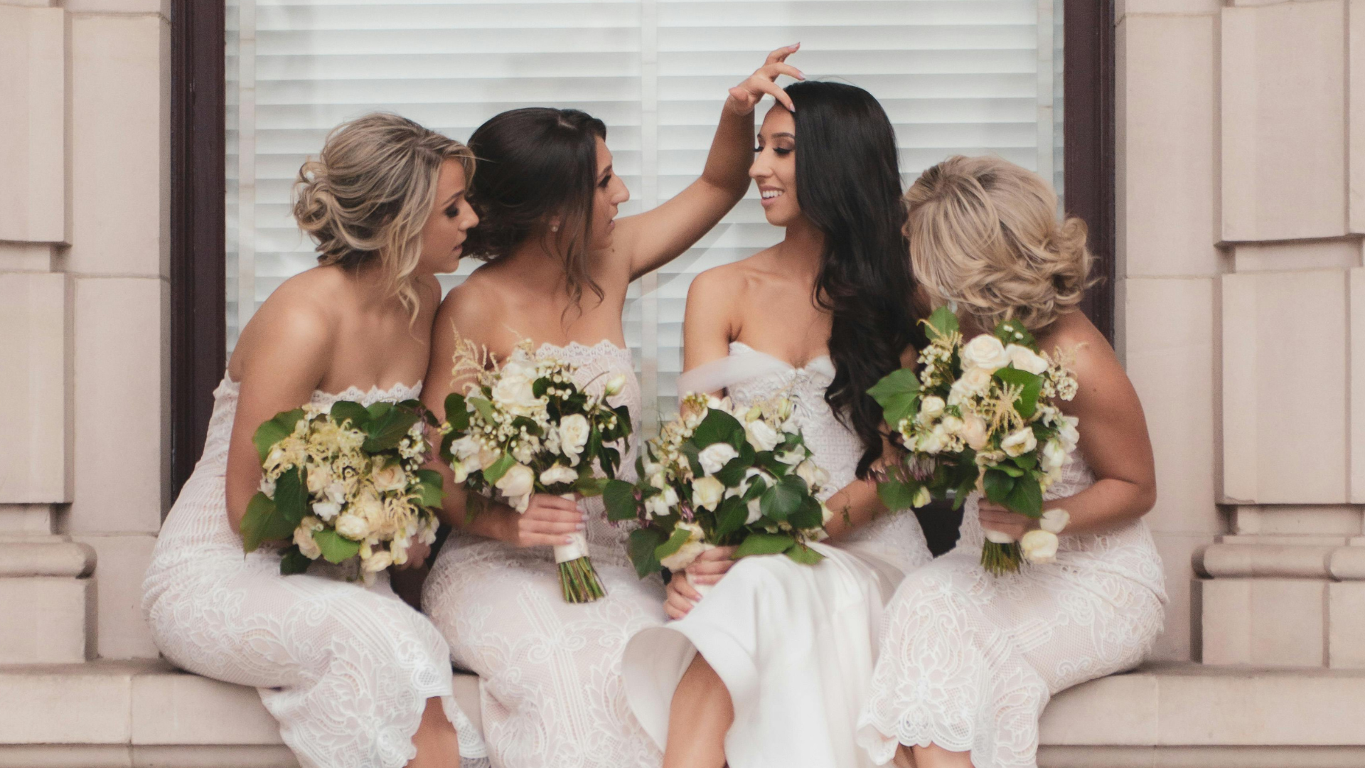 A bride and her bridesmaids are sitting on a window sill holding bouquets of flowers.