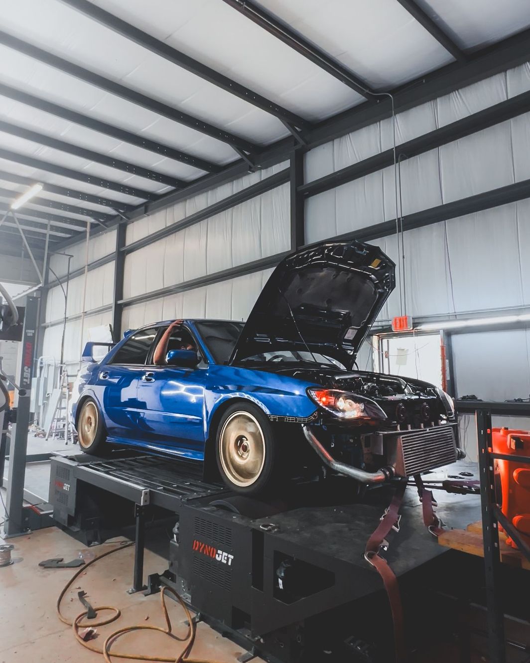 A blue Subaru WRX STI with its hood open, secured on a dynamometer inside a garage for testing.