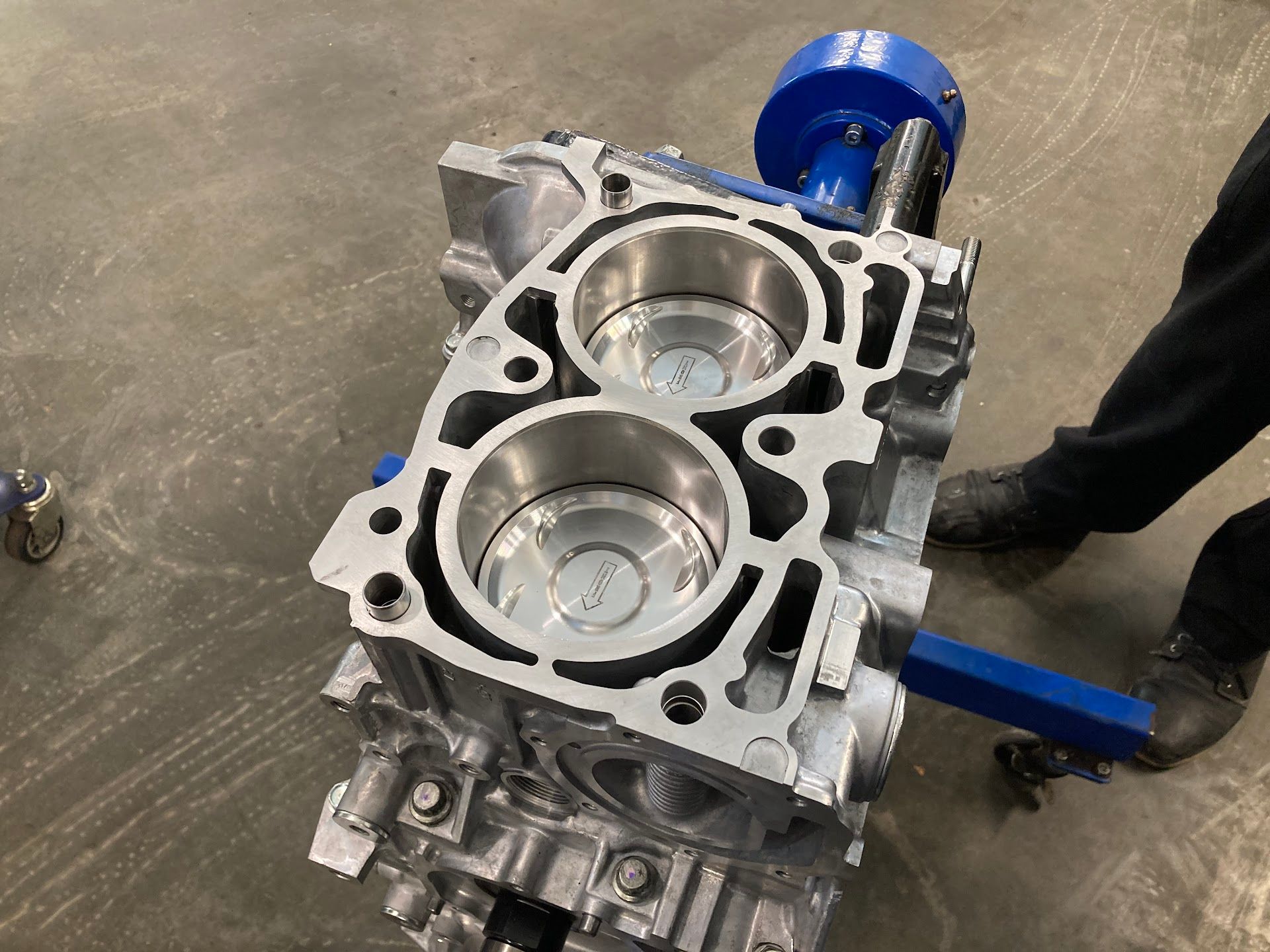 An aluminum engine block mounted on a blue stand in a workshop, showing two exposed cylinder bores. | Teter Auto