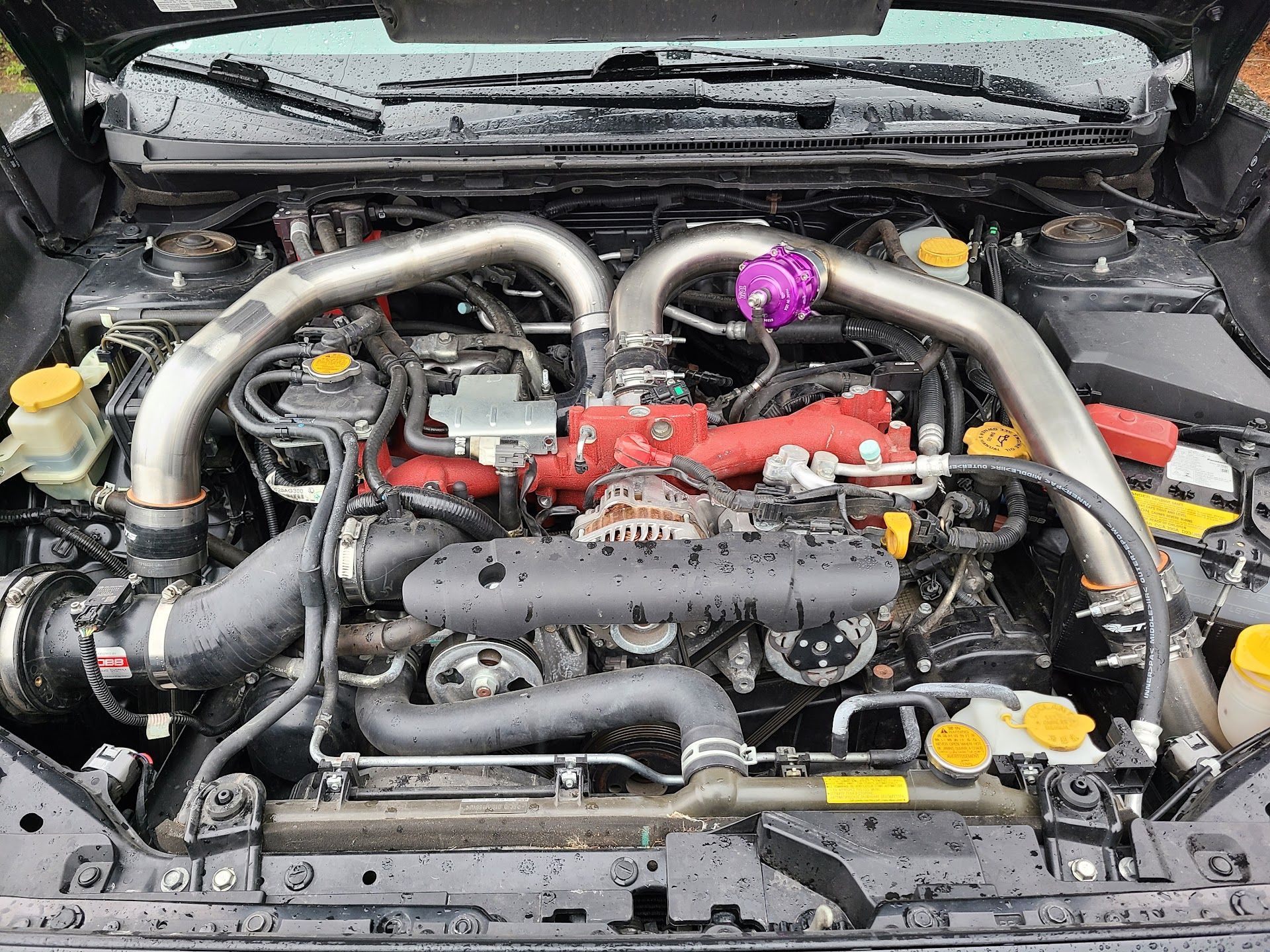 An open car hood showing a Subaru  engine bay with a prominent red intake manifold, metallic piping, and a purple part. | Teter Auto