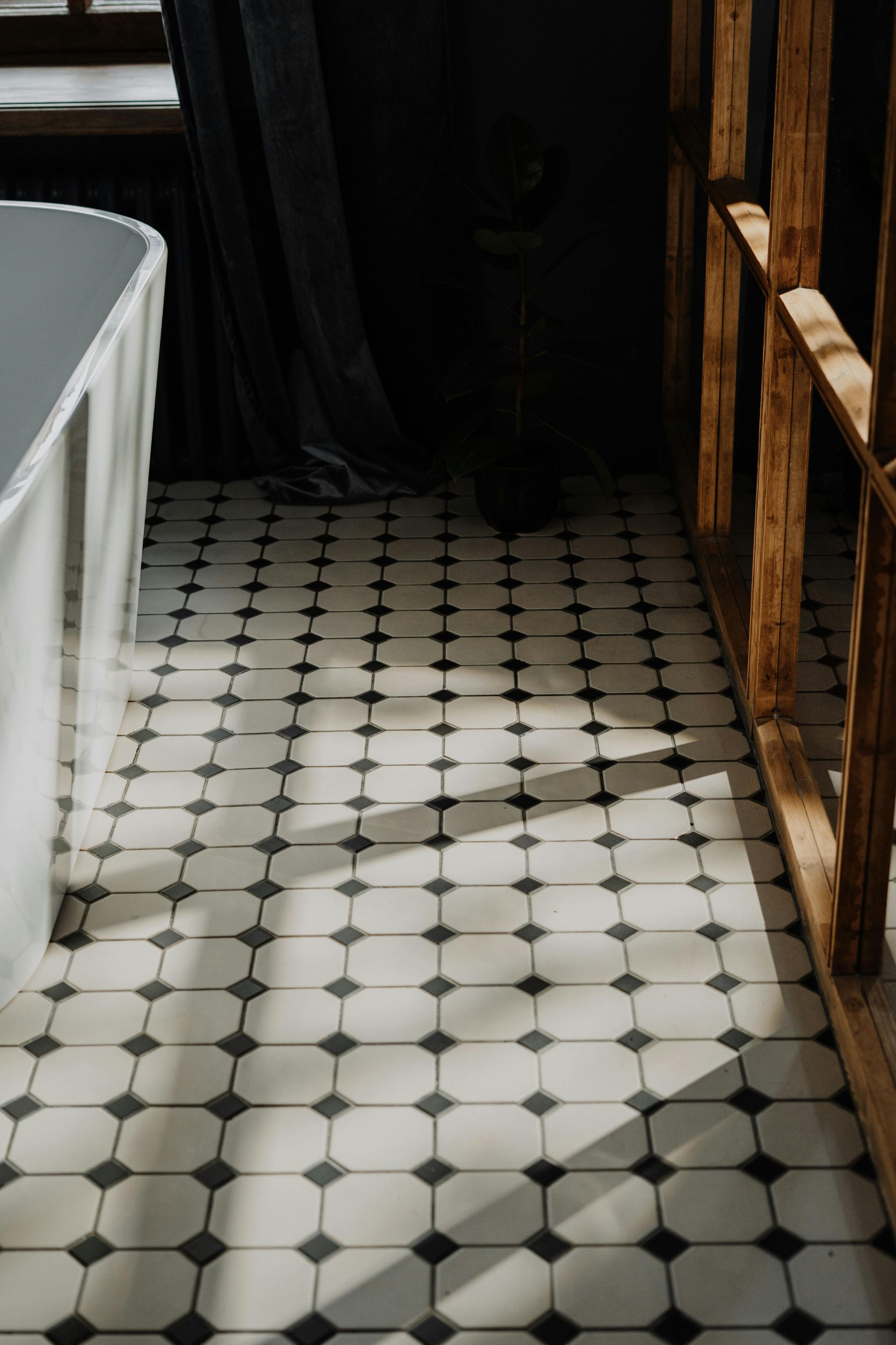 Sunlit black-and-white tiled floor beside a white bathtub and wooden railing