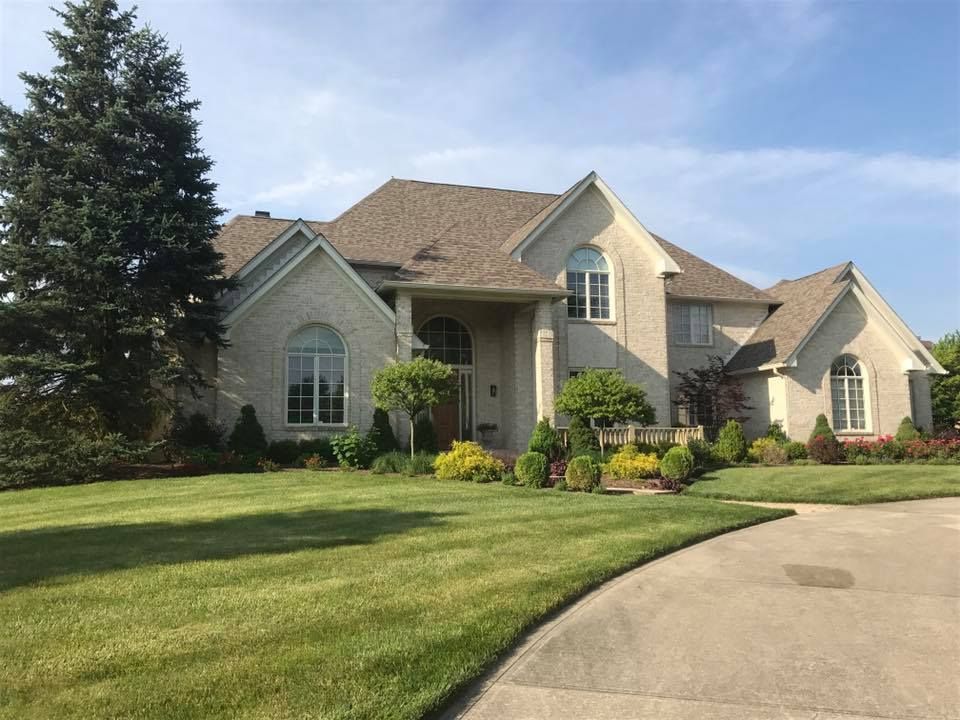 Beige brick two-story house with brown roof and curved driveway, surrounded by green grass and trees.