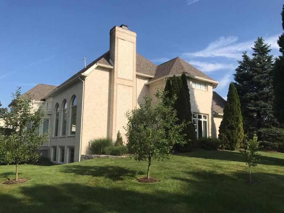 Beige house with a brown roof, chimney, and trees on a grassy lawn under a blue sky.