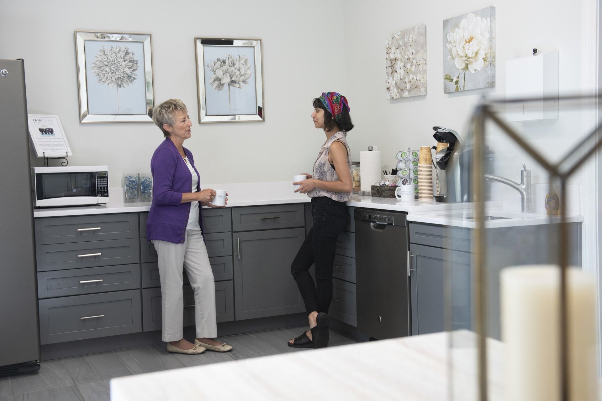 Two people converse in a bright kitchen. One wears a purple jacket and the other a patterned top. Gray cabinets, artwork, and appliances are visible.