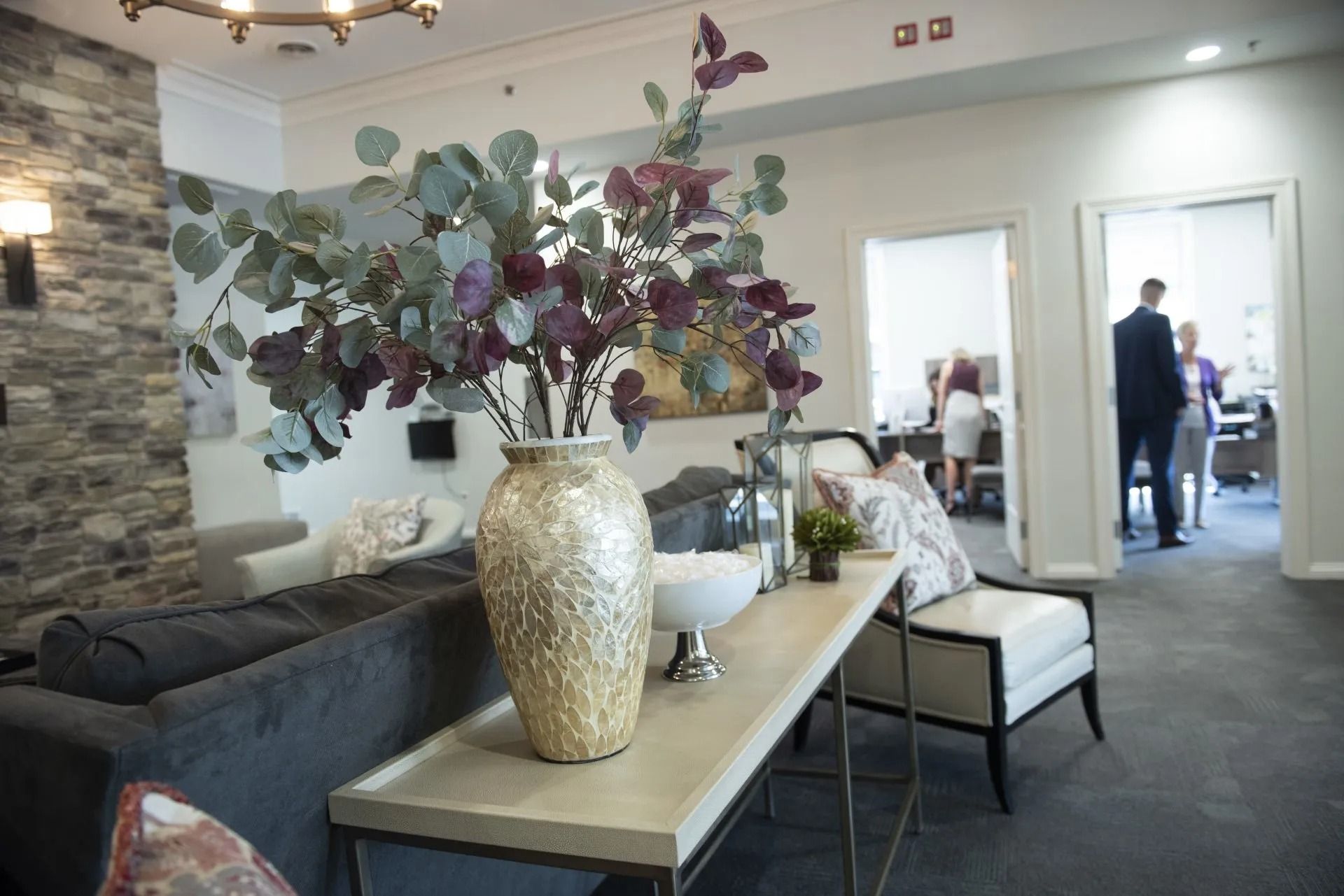 Elegant waiting room with grey sofa, console table, floral arrangement, and open doors with people.