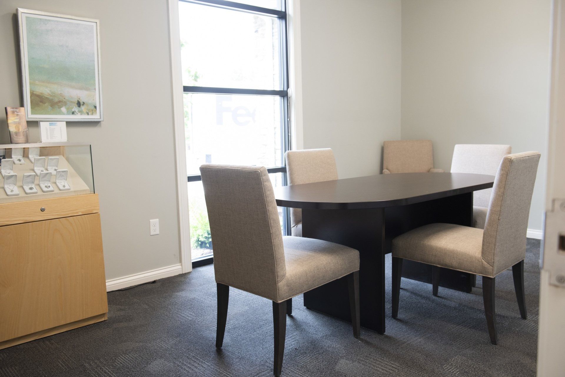 A small meeting room with a dark table and four chairs, a display case, and a window.