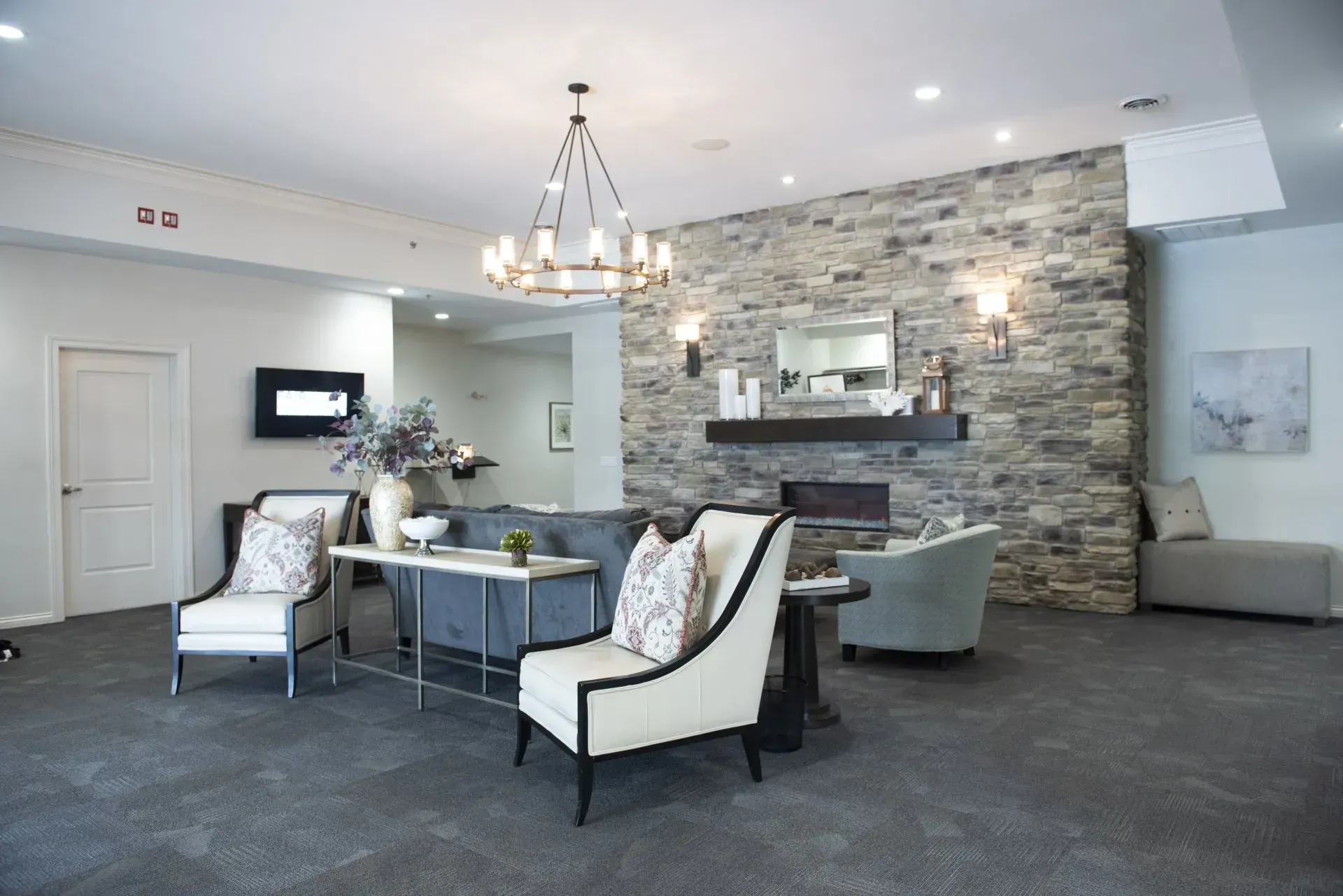 Lounge area with stone fireplace, sofas, armchairs, chandelier, and gray carpet.