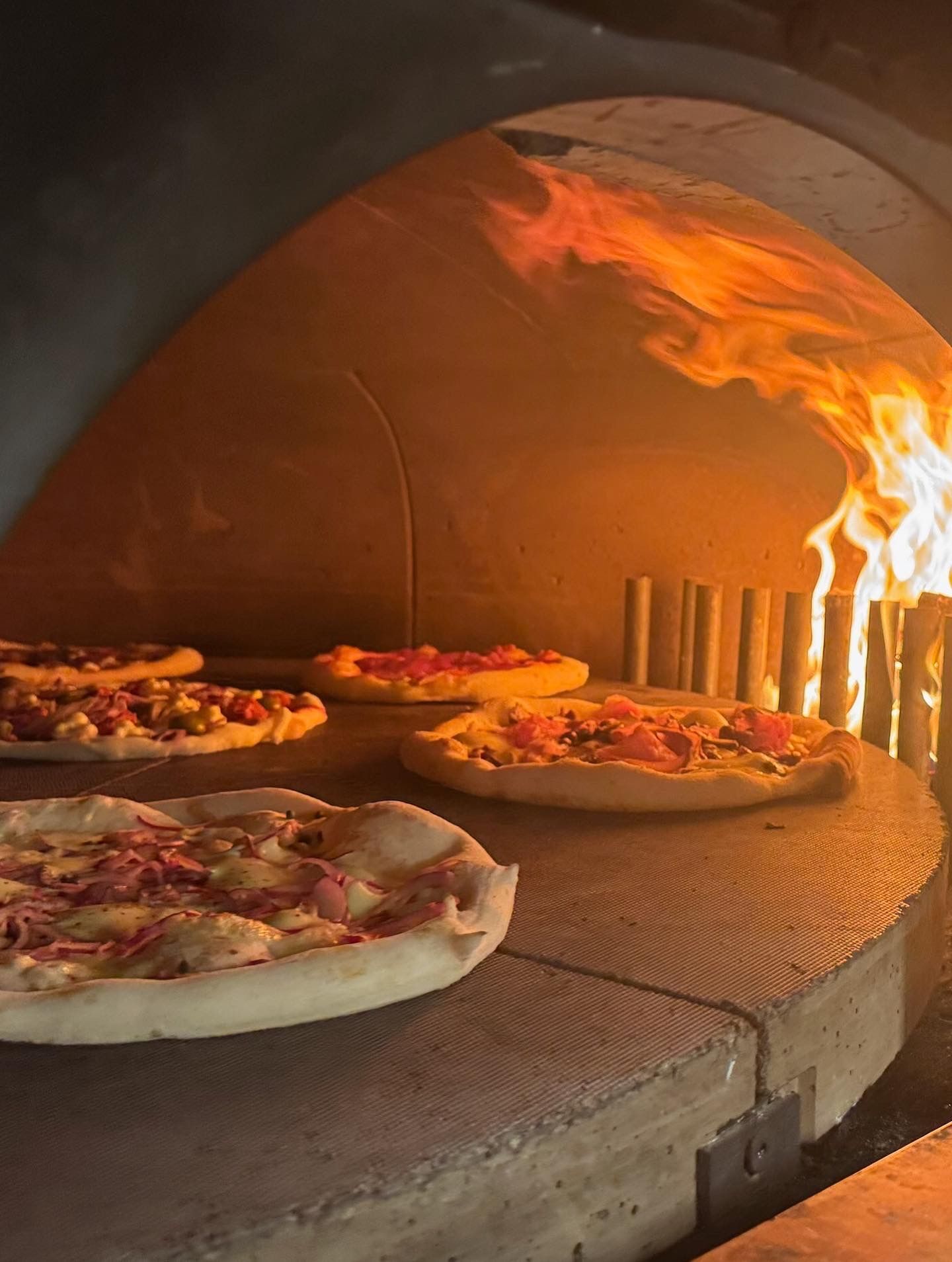 Pizzas cooking in a wood-fired oven with flames, in a warm, dimly lit environment.