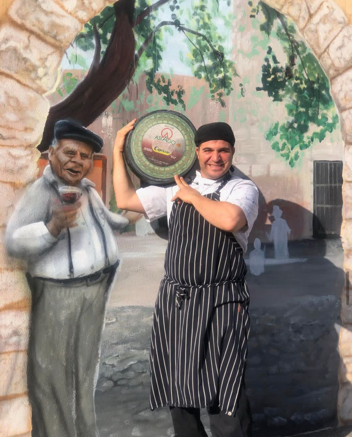 Chef holding a platter, posing with a painted mural of a man in Italy.