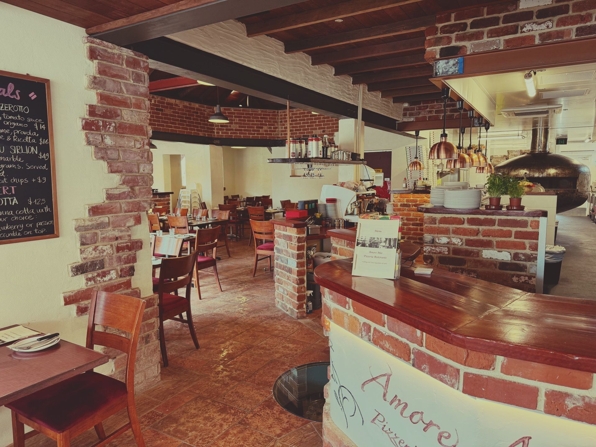 Interior of a brick-walled restaurant with tables and chairs, a pizza oven, and a bar.