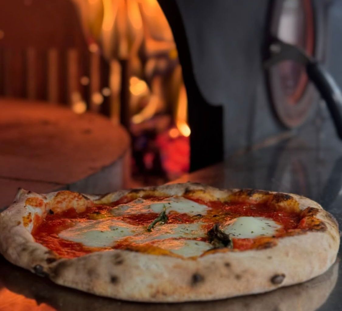 Pizza with mozzarella, tomato sauce, and basil in front of a wood-fired oven.