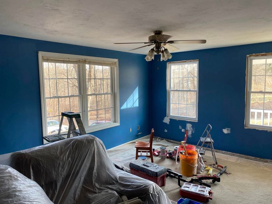 A room under renovation with bright blue walls, two large windows, a ceiling fan, and painting supplies scattered about.