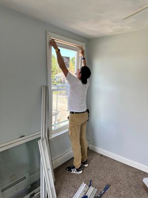A person wearing a white shirt and tan work pants installs a window frame in a room with blue walls and textured carpet.
