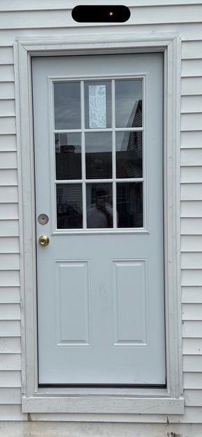 A white, nine-pane glass panel door installed in light gray vinyl siding with a brass-colored round doorknob.