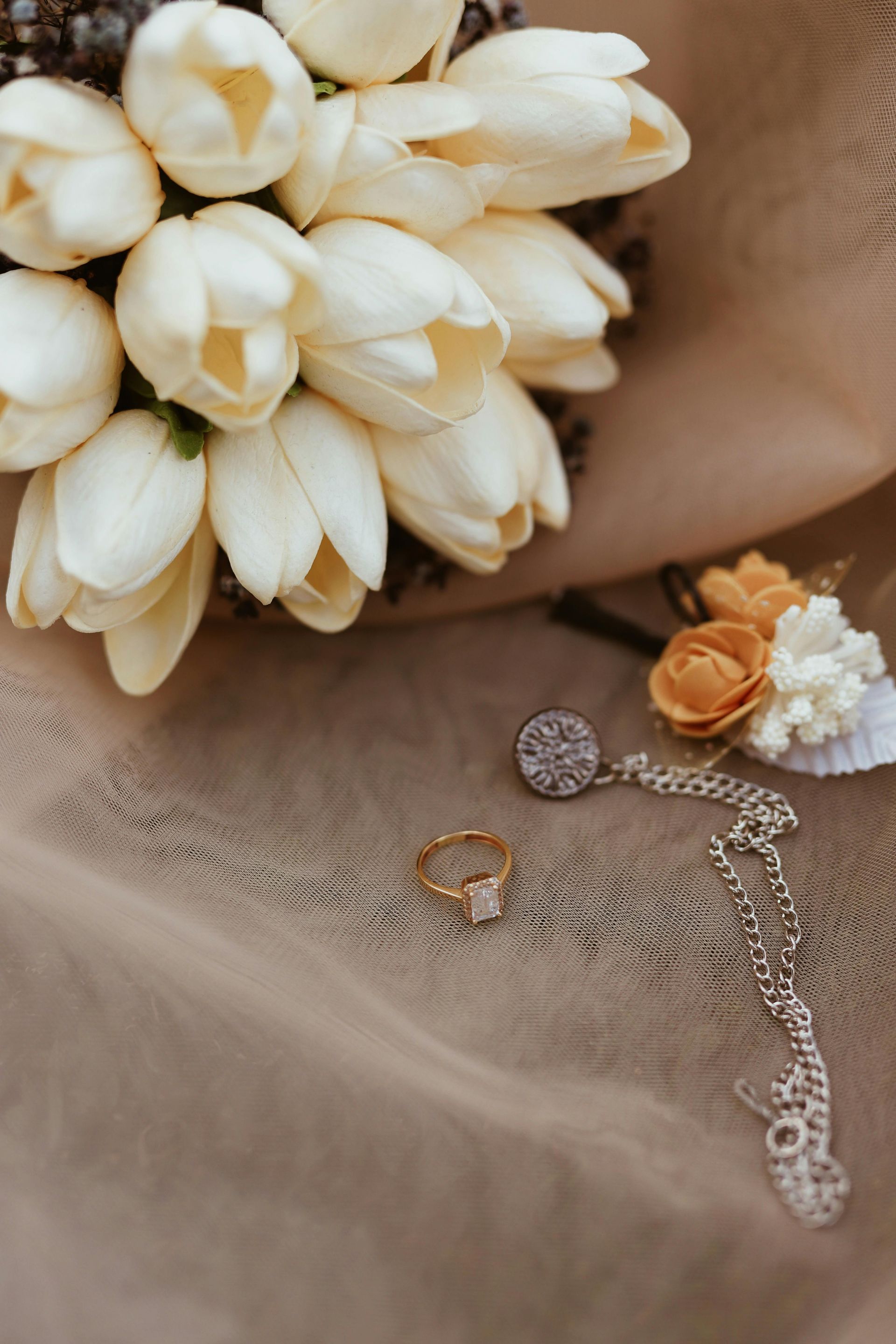 A wedding bouquet of white tulips rests on beige fabric next to a gold ring and a silver necklace