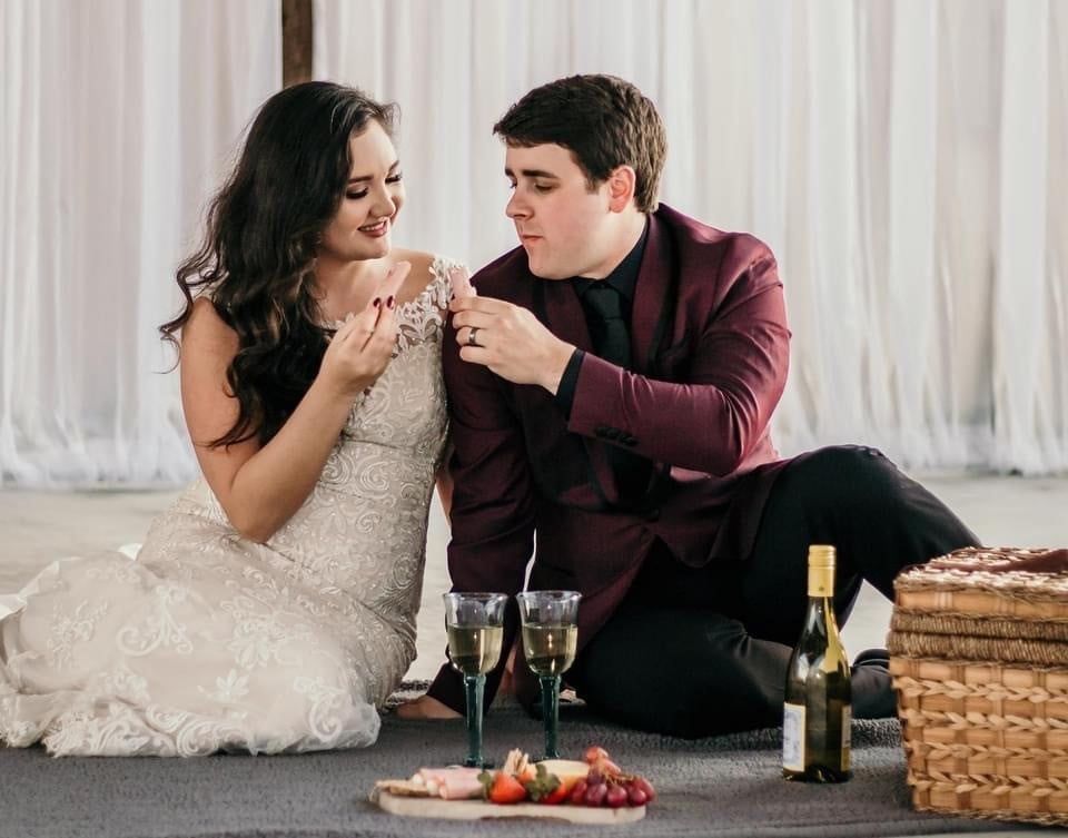 A bride and groom are sitting on the floor with wine glasses and food.
