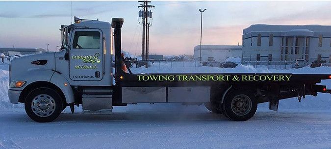 A towing truck is parked in the snow in front of a building.