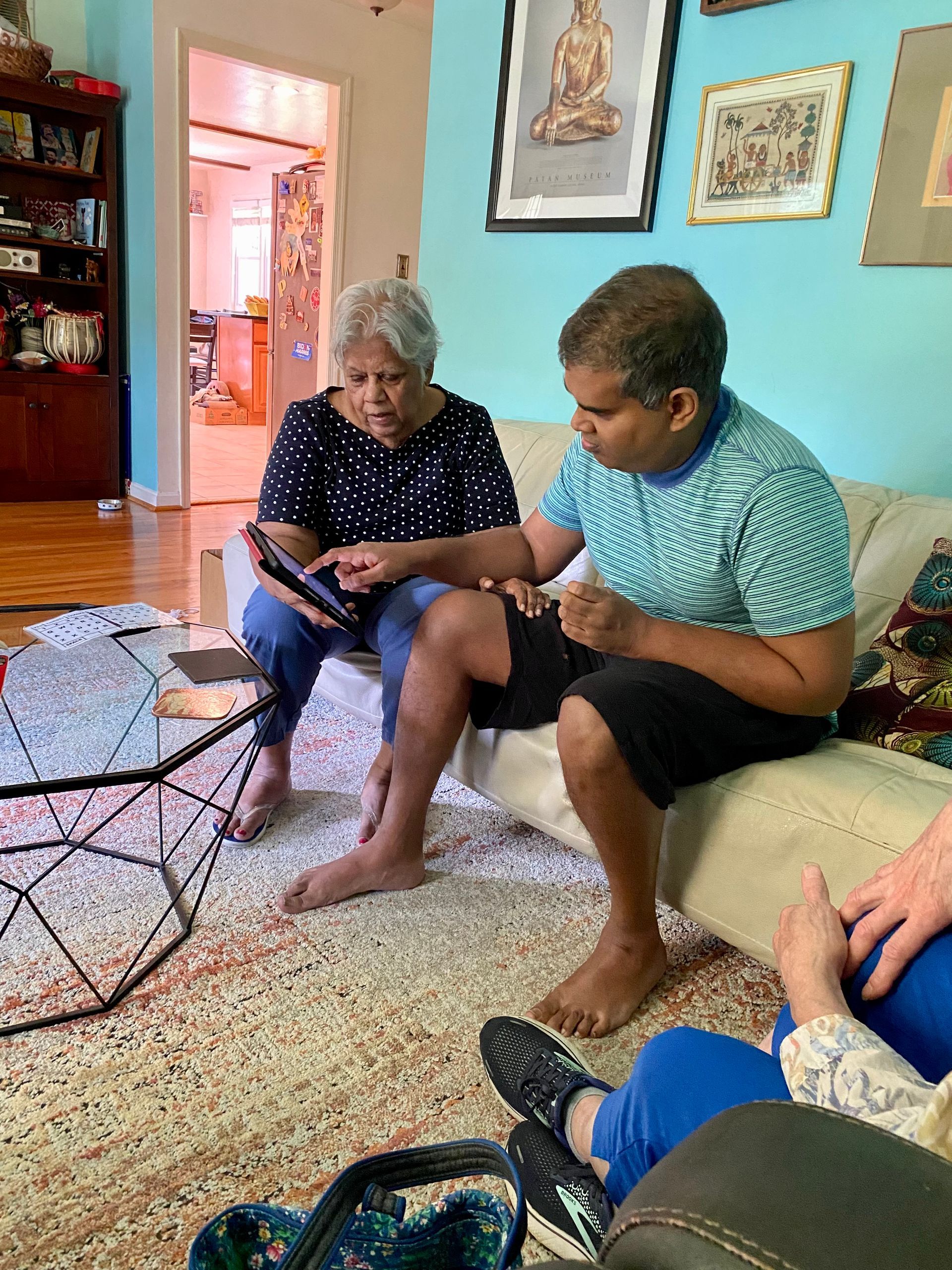 Two people looking at a tablet on a couch; light blue wall, patterned rug, other person's legs visible.