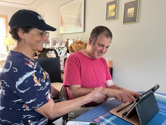 Woman in cap helps man with tablet; both smile indoors.