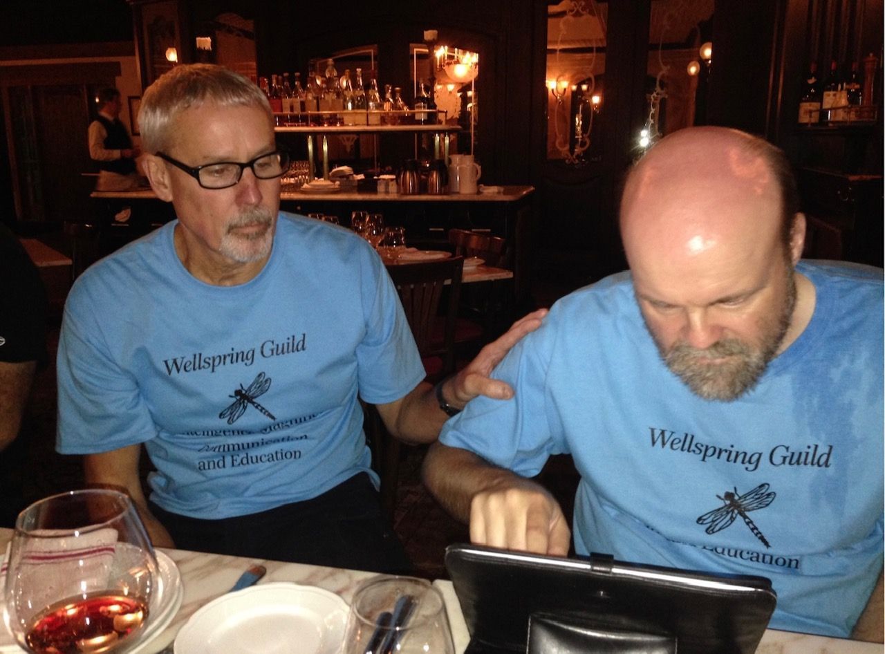 Two men in matching blue shirts at a restaurant. One touches the other's shoulder, both looking at a tablet.