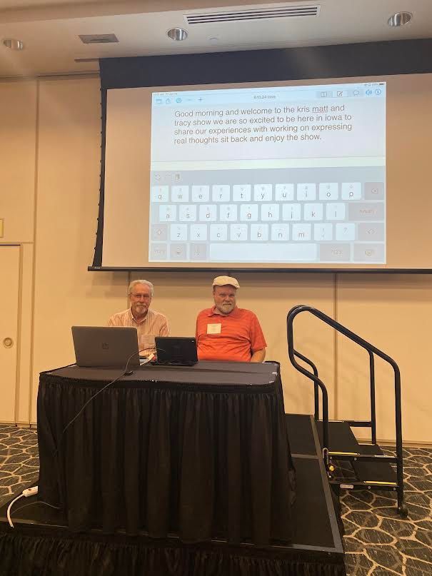 Two men presenting at a conference, using laptops and a projection screen showing text and a keyboard.