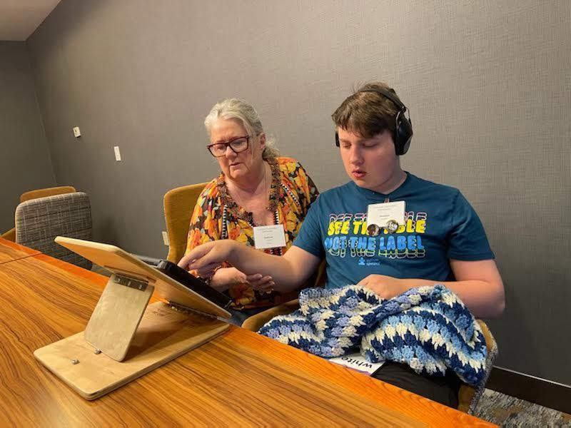 Woman and boy using a laptop at a table. The boy wears headphones and looks at the screen. They appear focused.