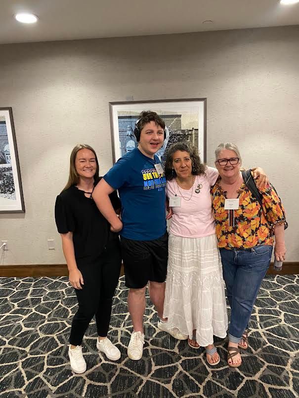 Four people pose together. Two women and two people with smiles. The group is indoors with a carpet.
