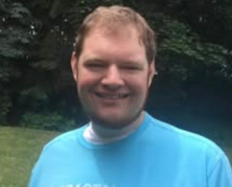 Man with light skin and blond hair smiles in a blue shirt outside on a green lawn.