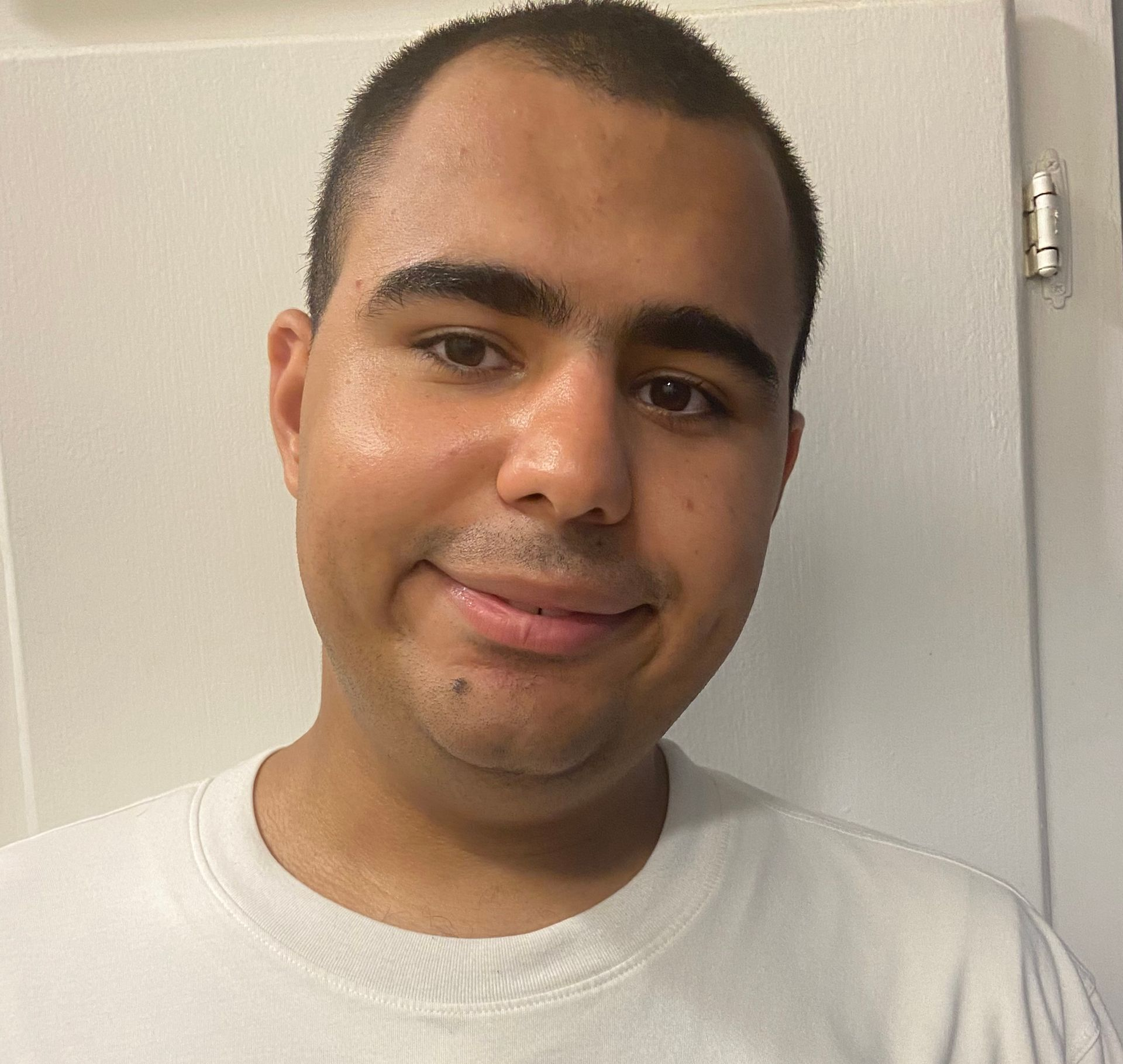 Man with short dark hair and a light complexion smiling, wearing a white t-shirt, indoors.