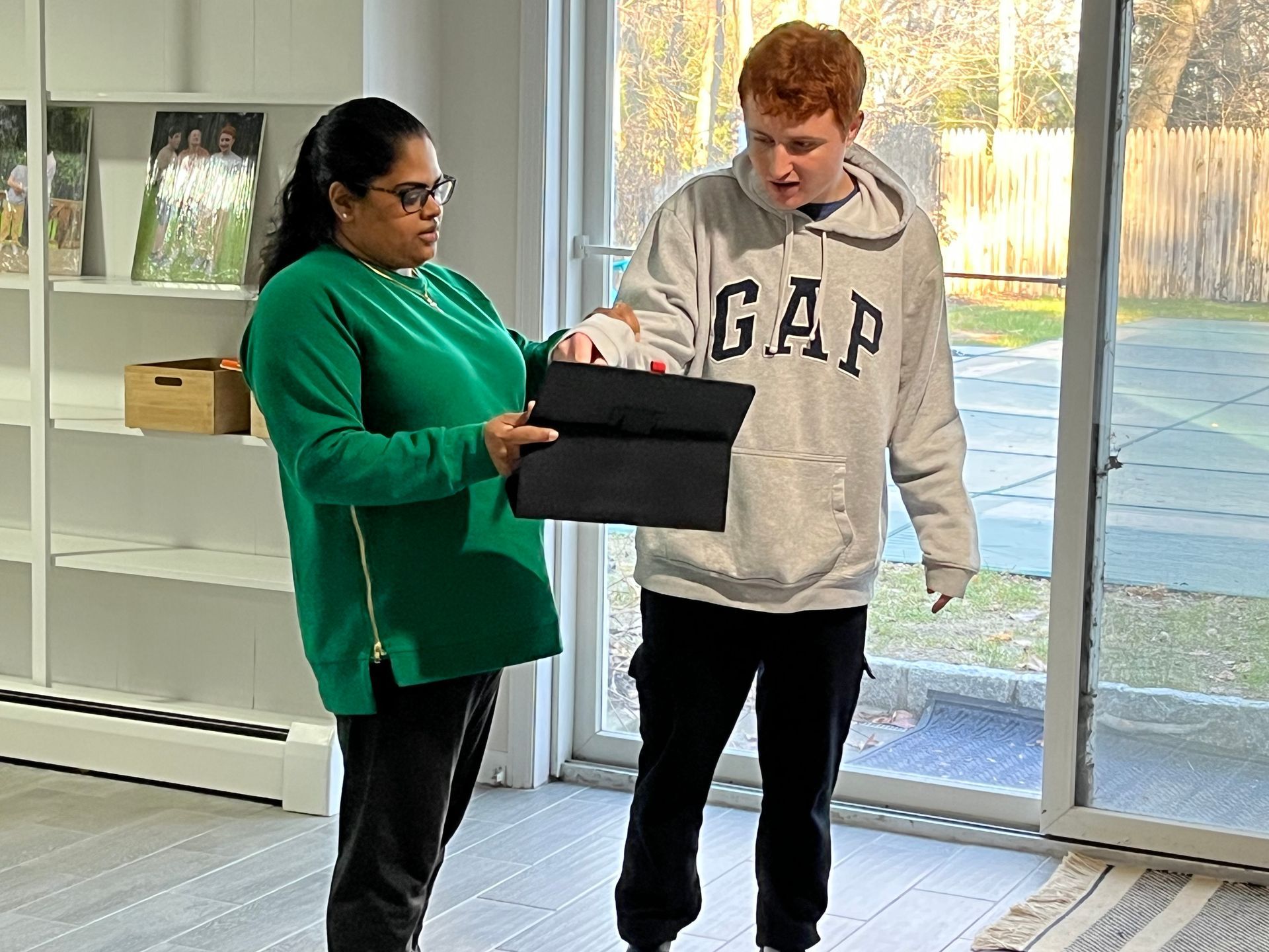 Woman in green shirt shows a folded black item to a young man in a GAP hoodie inside a bright room.