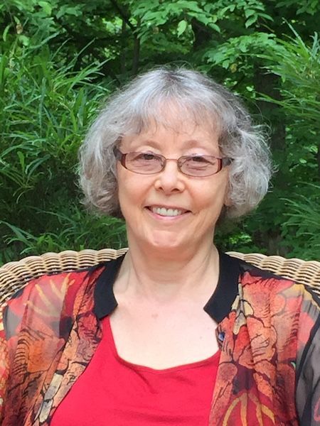 Woman with glasses smiles; red top, patterned jacket, grey hair, outdoor setting.