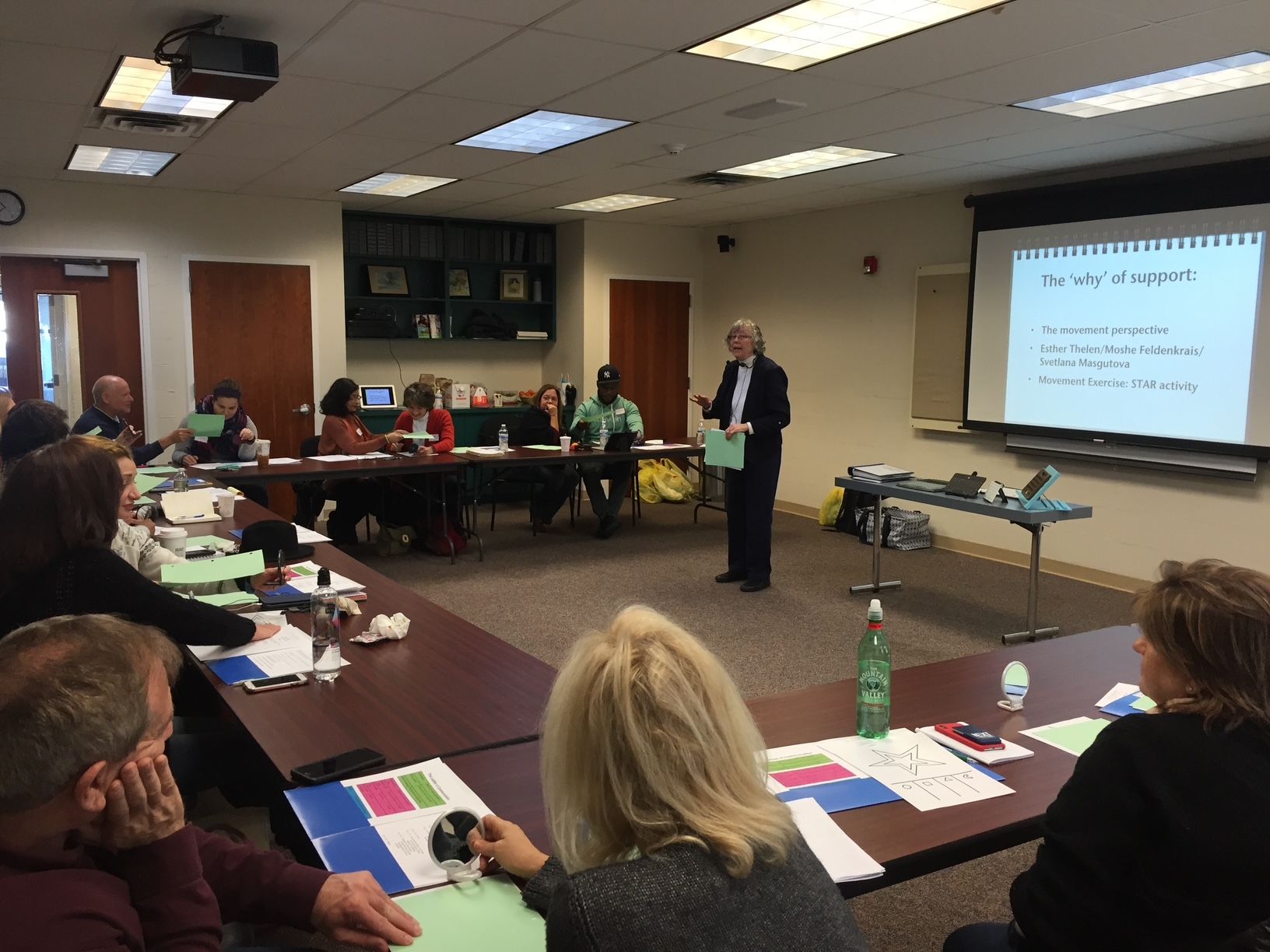 People in a meeting with a presenter. A projection screen shows text.