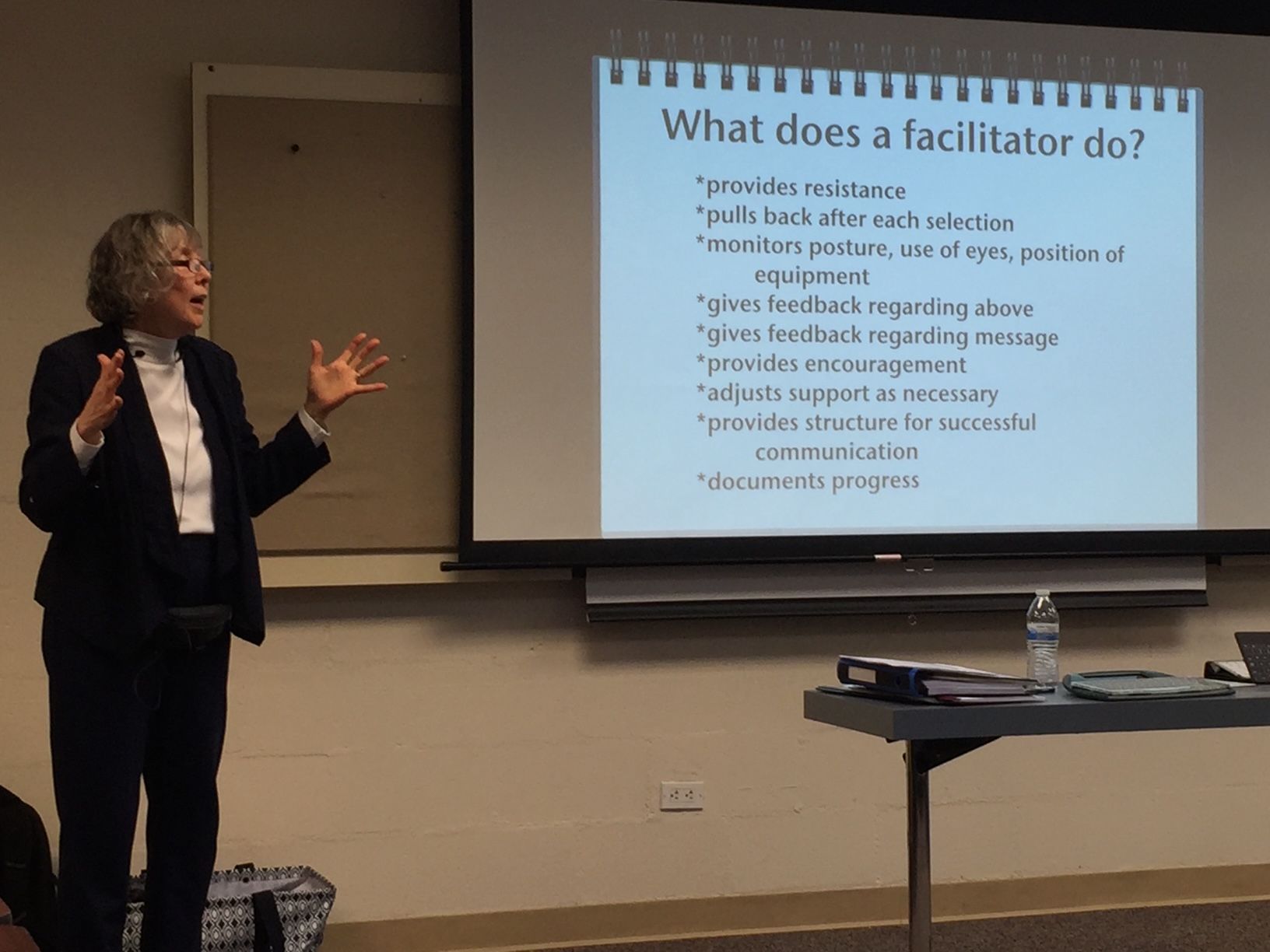 A woman presents in front of a screen listing facilitator duties. She gestures, standing near a table with materials.
